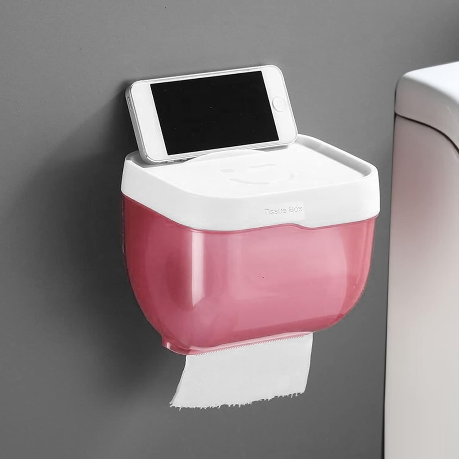 White bathroom tissue dispenser with a phone resting on its top shelf, showing its self-adhesive installation.
